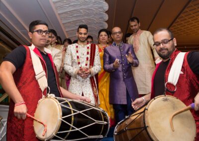 Two drummers play traditional instruments as a groom and guests watch intently.