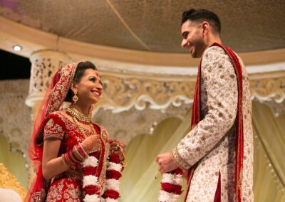 A bride and groom smile at each other, holding garlands during a ceremony.