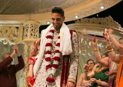A groom smiles, wearing a white and gold outfit with red and white garlands.