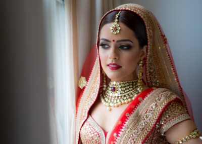 Bride looking out window in red and gold lehenga before wedding.