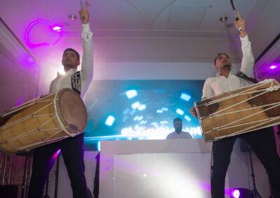 Two dhol players performing energetically on stage with DJ and lights behind.