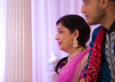 Smiling woman in pink saree watching wedding rituals beside man in blue traditional attire.