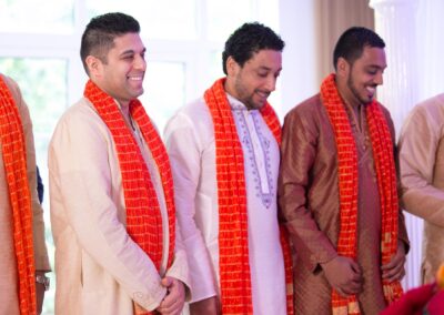 Groomsmen smiling during wedding ceremony wearing cream kurtas and orange scarves.