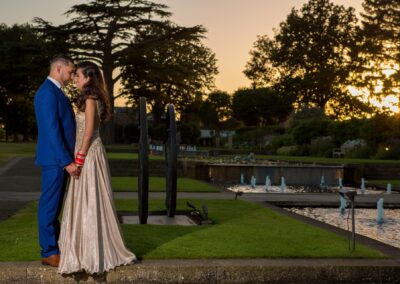 Bride and groom holding hands at sunset in garden with fountain and elegant greenery.