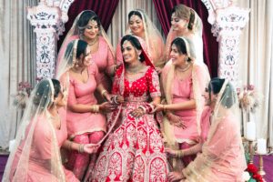 A bride in a red lehenga surrounded by bridesmaids in pink dresses.