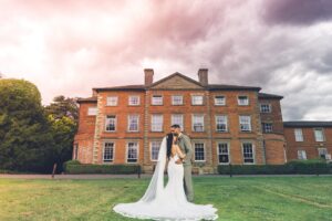 A bride and groom embrace on a lawn in front of a large brick mansion.