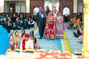 A bride walks down an aisle in a grand hall, surrounded by wedding guests.