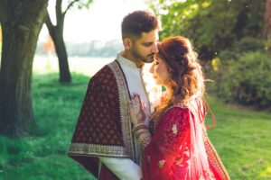 A groom gently kisses his bride's forehead in a sunlit outdoor setting.