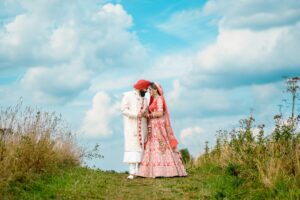 A bride and groom in traditional attire pose under a cloudy sky in a field.