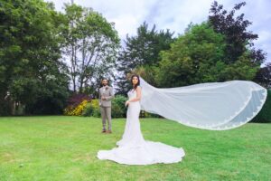 A bride in a white gown with a flowing veil and a groom in a suit standing in a garden.