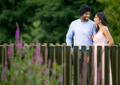 Couple standing together on wooden bridge surrounded by greenery and flowers.