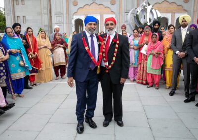 Two elders wearing garlands posing together outside Gurdwara surrounded by family members.