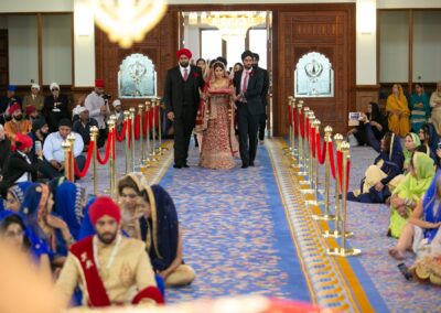 Bride entering Gurdwara aisle escorted by family members for Sikh wedding ceremony.