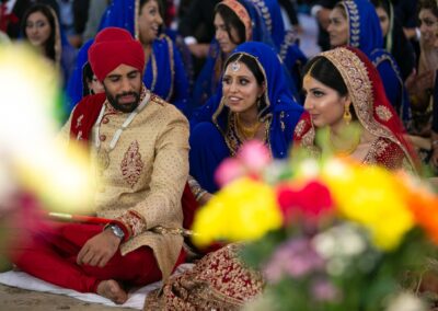 Bride and groom smiling during Sikh wedding ceremony surrounded by guests in blue outfits.