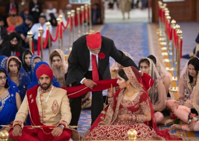 Sikh wedding ceremony with elder tying red scarf between bride and groom during Anand Karaj.