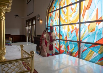 Bride and groom walking beside stained glass window inside Gurdwara.