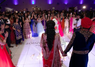 Couple standing before wedding cake as guests cheer during cake-cutting moment.