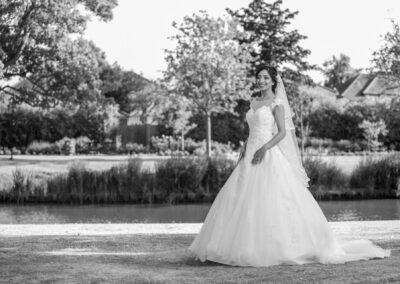 Bride in white lace gown smiling beside river during outdoor wedding portrait.