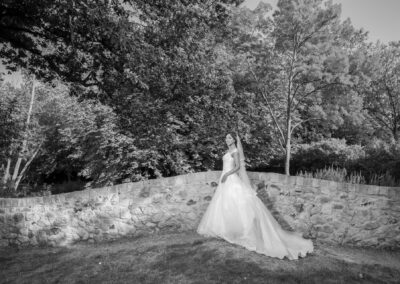 Bride in white gown posing outdoors by stone wall under sunlight.